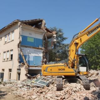 Escavatore demolisce edificio industriale sotto un cielo sereno.
